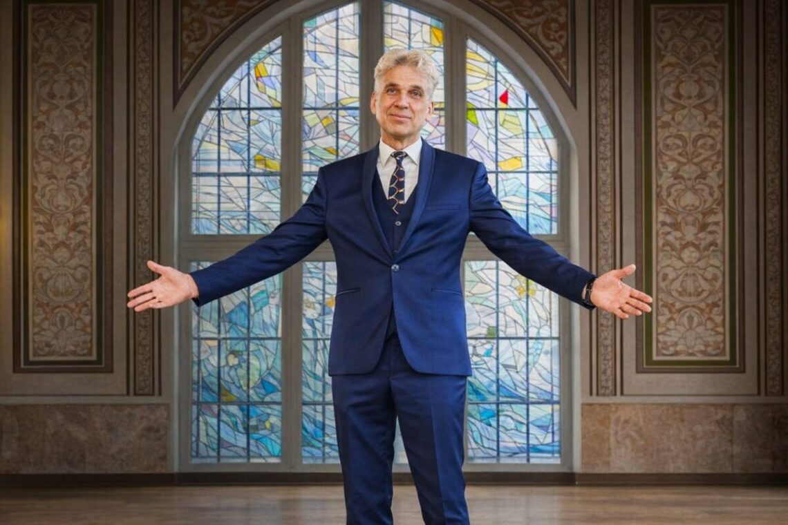 Rimantas Balsys išskiestomis rankomis Klaipėdos universiteto Vitražų menės fone