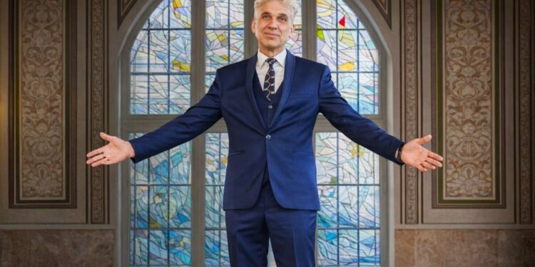 Rimantas Balsys išskiestomis rankomis Klaipėdos universiteto Vitražų menės fone