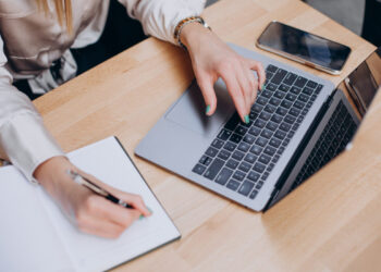 Nešiojamas kompiuteris, popieriaus lapas ir rašanti ranka