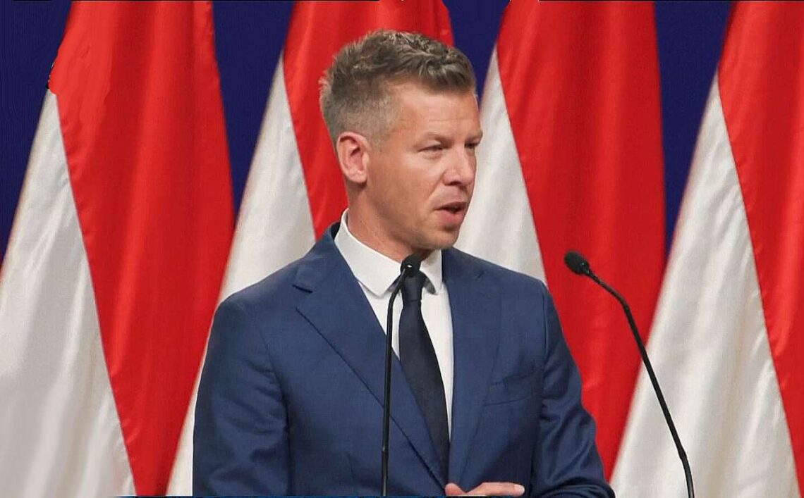 Pėteris Madjaras kalba prie tribūnos su šalies vėliavomis fone