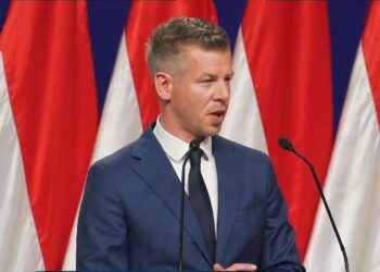 Pėteris Madjaras kalba prie tribūnos su šalies vėliavomis fone