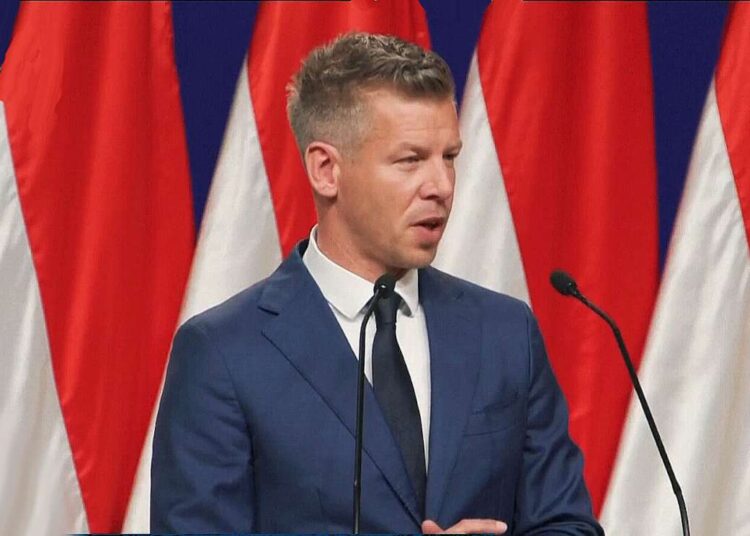 Pėteris Madjaras kalba prie tribūnos su šalies vėliavomis fone