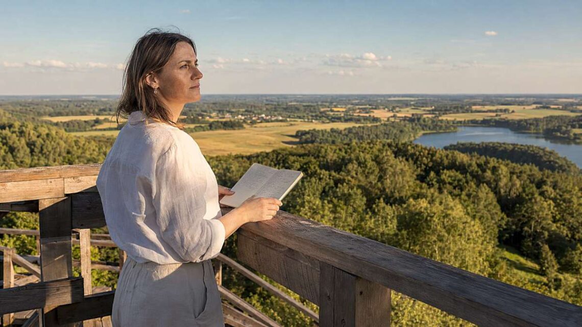 Moteris apžvalgos aikštelėje su užrašų knygele žvelgia į vasarišką Lietuvos kraštovaizdį