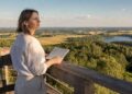 Moteris apžvalgos aikštelėje su užrašų knygele žvelgia į vasarišką Lietuvos kraštovaizdį