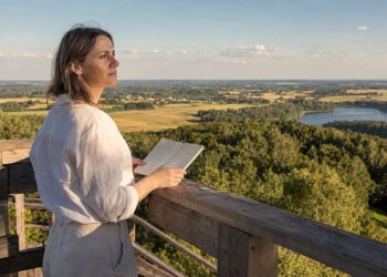 Moteris apžvalgos aikštelėje su užrašų knygele žvelgia į vasarišką Lietuvos kraštovaizdį