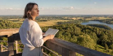 Moteris apžvalgos aikštelėje su užrašų knygele žvelgia į vasarišką Lietuvos kraštovaizdį