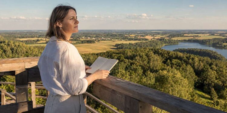 Moteris apžvalgos aikštelėje su užrašų knygele žvelgia į vasarišką Lietuvos kraštovaizdį