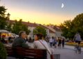 Du žmonės ramiai kalbasi ant suolelio Lietuvos senamiesčio aikštėje vasaros vakarą, virš miesto matomas jaunas dylantis Mėnulis.