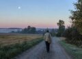 Žmogus ankstyvą rugsėjo rytą eina Lietuvos kaimo keliu, danguje matyti dylantis Mėnulis