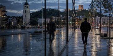 Žmogus vakare eina lietaus nuplauta Vilniaus gatve, miesto šviesoms atsispindint stikle