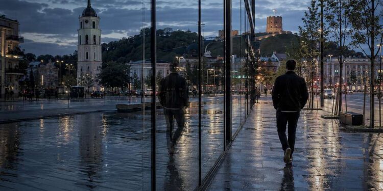 Žmogus vakare eina lietaus nuplauta Vilniaus gatve, miesto šviesoms atsispindint stikle