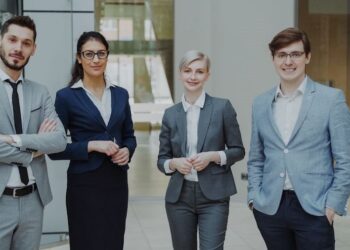 Keturi biuro darbuotojai stovi šiuolaikiškoje darbo aplinkoje