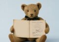 Pliušinis meškiukas laiko atverstą 1933 m. knygos „Winnie-the-Pooh“ leidimą su Abelio Balošerio bibliotekos antspaudu.