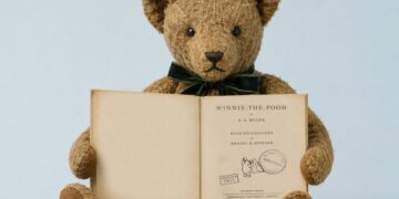 Pliušinis meškiukas laiko atverstą 1933 m. knygos „Winnie-the-Pooh“ leidimą su Abelio Balošerio bibliotekos antspaudu.