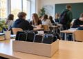 Mokyklos klasėje specialioje dėžėje sudėti mokinių mobilieji telefonai, fone bendrauja mokiniai