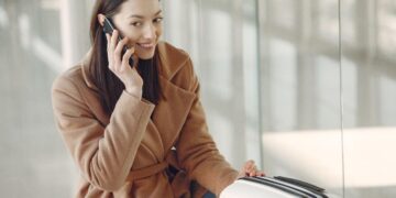 Moteris rudu paltu kalba mobiliuoju telefonu, šalia jos baltas kelioninis lagaminas